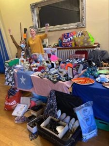 Table set up with various items including jewellery, bags, ornaments, books. Faye and Sarah stood behind the table with arms raised in celebration.