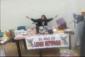 Faye is sat behind the table, she is smiling and wearing a black leigh network t-shirt, she has her arms spread wide showing off the stall table which has lots of different items for sale on.